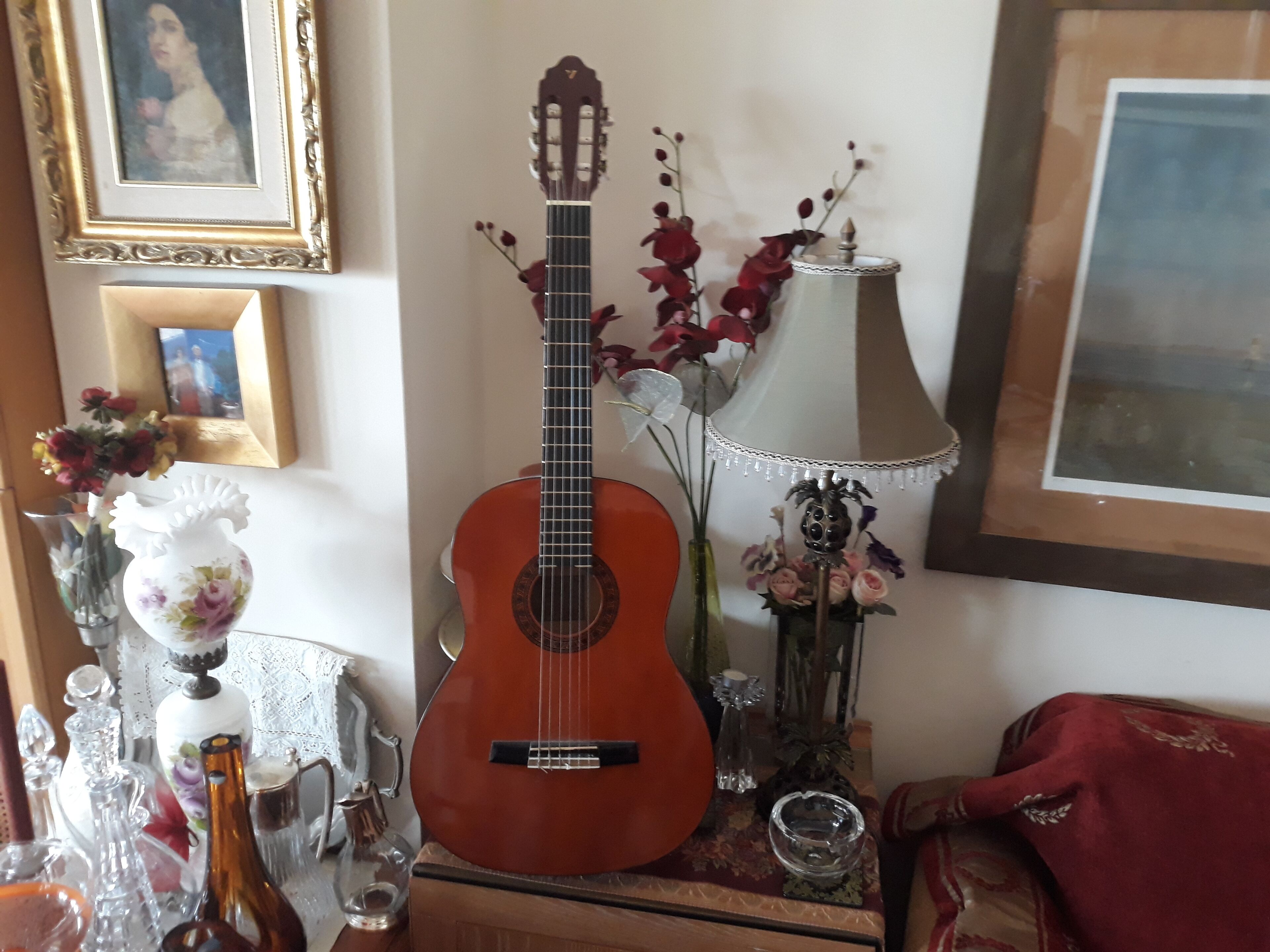 Une guitare posée sur une surface en bois, entourée d’objets décoratifs, notamment un vase avec des fleurs, une lampe et un tableau encadré accroché au mur.