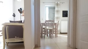 A modern interior with a dining area featuring a white table and chairs, a small desk with a lamp, and a signboard. - New, large apartm. with private garden, 3' to the sea and 5' to the city centre (Καλαμάτα)