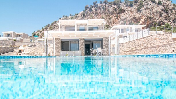 A swimming pool with a stone wall and a building with a balcony in the background. - Gregorys Luxury Villa in Kallithea (Αφάντου)