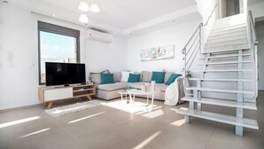 A modern living room with a flat-screen TV, a sofa with teal cushions, a white coffee table, and a staircase. - Gregorys Luxury Villa in Kallithea (Αφάντου)