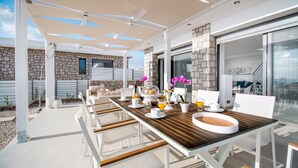 A sunlit outdoor dining area with a long table, white chairs, and a stone wall backdrop. - Gregorys Luxury Villa in Kallithea (Αφάντου)