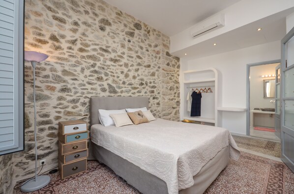A bedroom with a stone wall, a bed with a grey headboard, a bedside table with drawers, and a hanging coat rack.