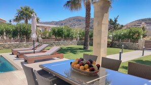 A poolside area with lounge chairs, a table with a fruit bowl, and a view of mountains. - Alexandros villa with private pool (Νωπήγεια)