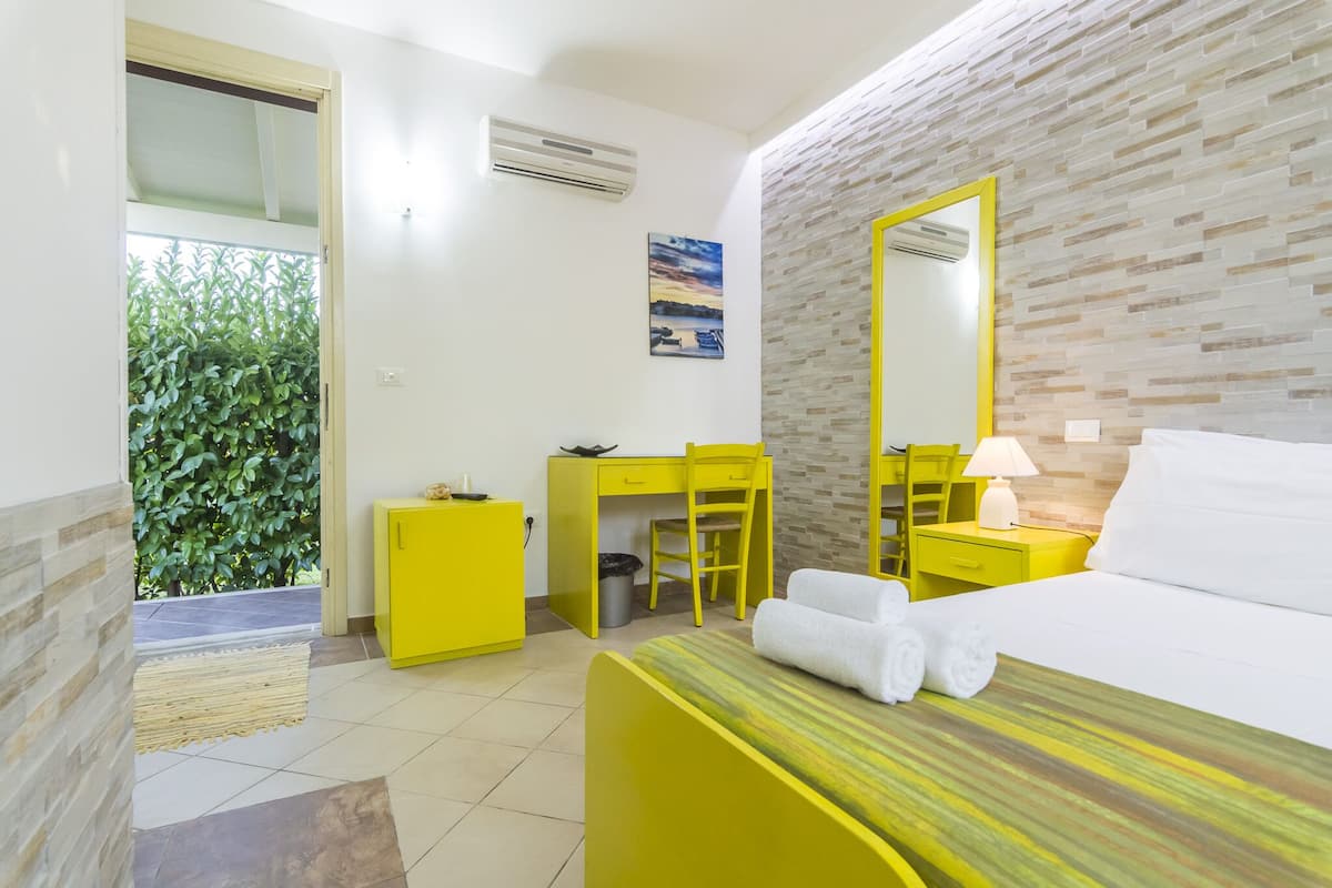 Habitación de hotel con cama, escritorio, silla, mueble pequeño, espejo y ventana con vista a follaje.