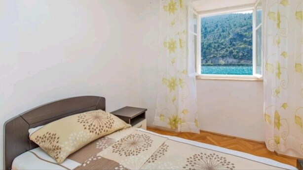 A bedroom with a bed, a nightstand, and a window with curtains. - Apartment Monkovic (Dubrovnik)