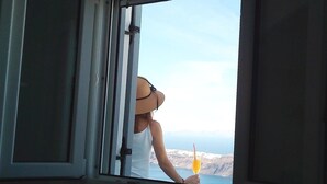 A person holding a drink by a window with a view of a rocky coastline. - Villa Renieris in Imerovigli, Santorini (Σαντορίνη)