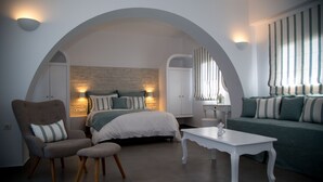 A modern bedroom with a bed, a sofa, a chair, and a table. There are two wall-mounted lights and a window with curtains. - Villa Renieris in Imerovigli, Santorini (Σαντορίνη)