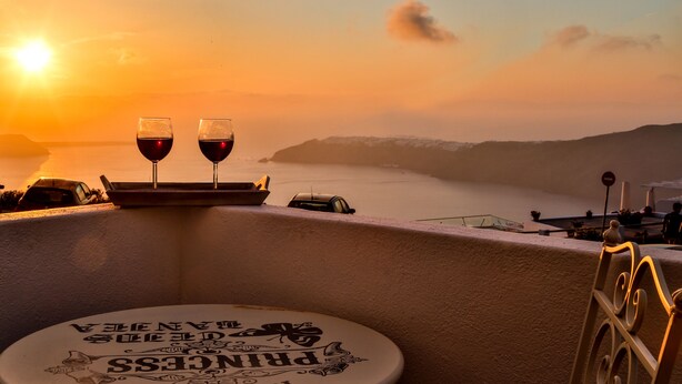 A table with two wine glasses and a sign on a balcony overlooking a sunset and water. - Villa Renieris in Imerovigli, Santorini (Σαντορίνη)