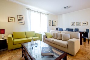 A living room with a green sofa, a beige sofa, and a glass coffee table with a book and decorative items. There are framed pictures on the walls and a dining area with a table and chairs.