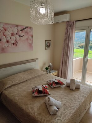 A bedroom with a bed, pillows, a nightstand, a lamp, and a view of greenery through a window.