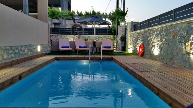 Una piscina rectangular con un muelle de madera alrededor. Se observan sillones de descanso y una mesa con sombrillas al fondo.