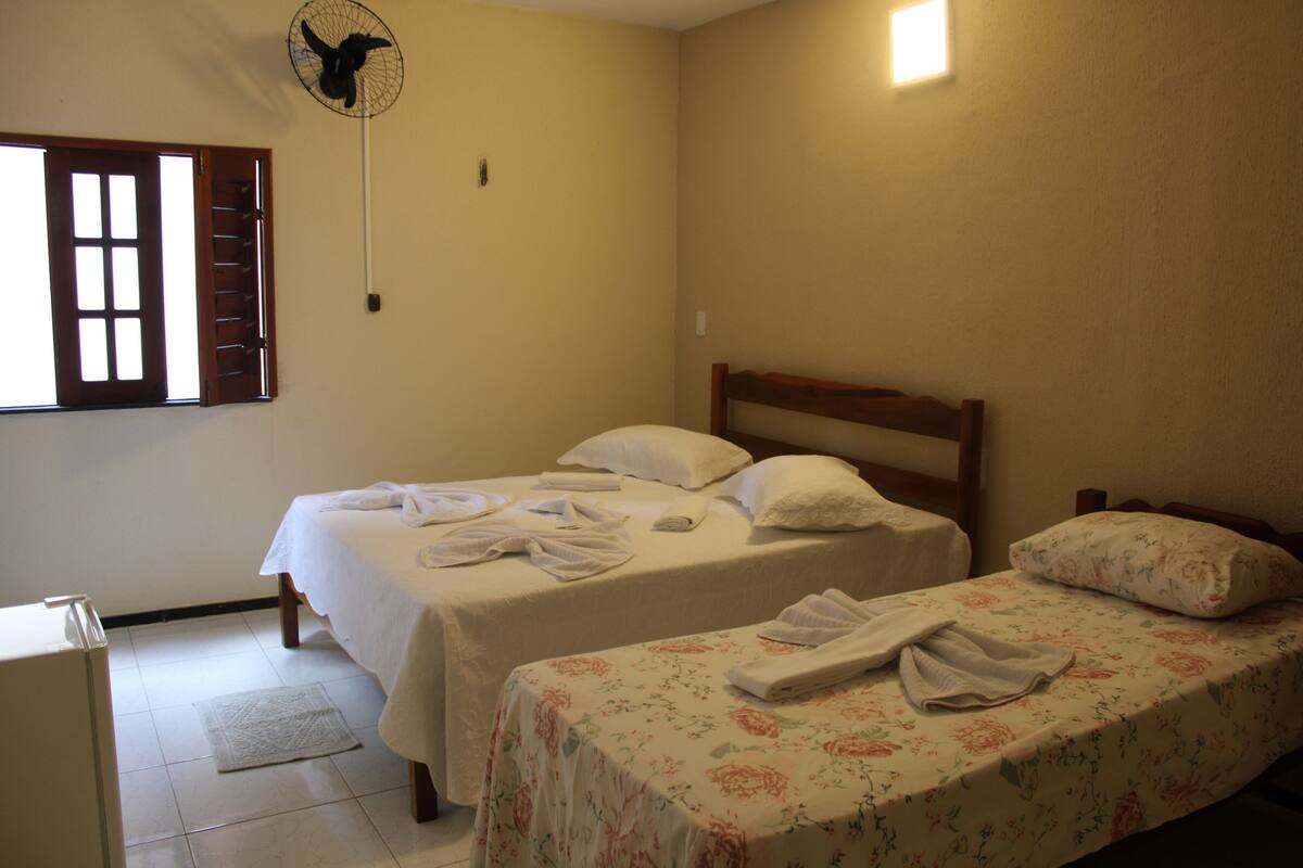 Une chambre avec deux lits, chacun recouvert d’une couvre-lit à motifs floraux et agrémenté d’oreillers blancs. On y trouve un ventilateur fixé au mur, une fenêtre avec des volets en bois et une petite table avec une lampe.
