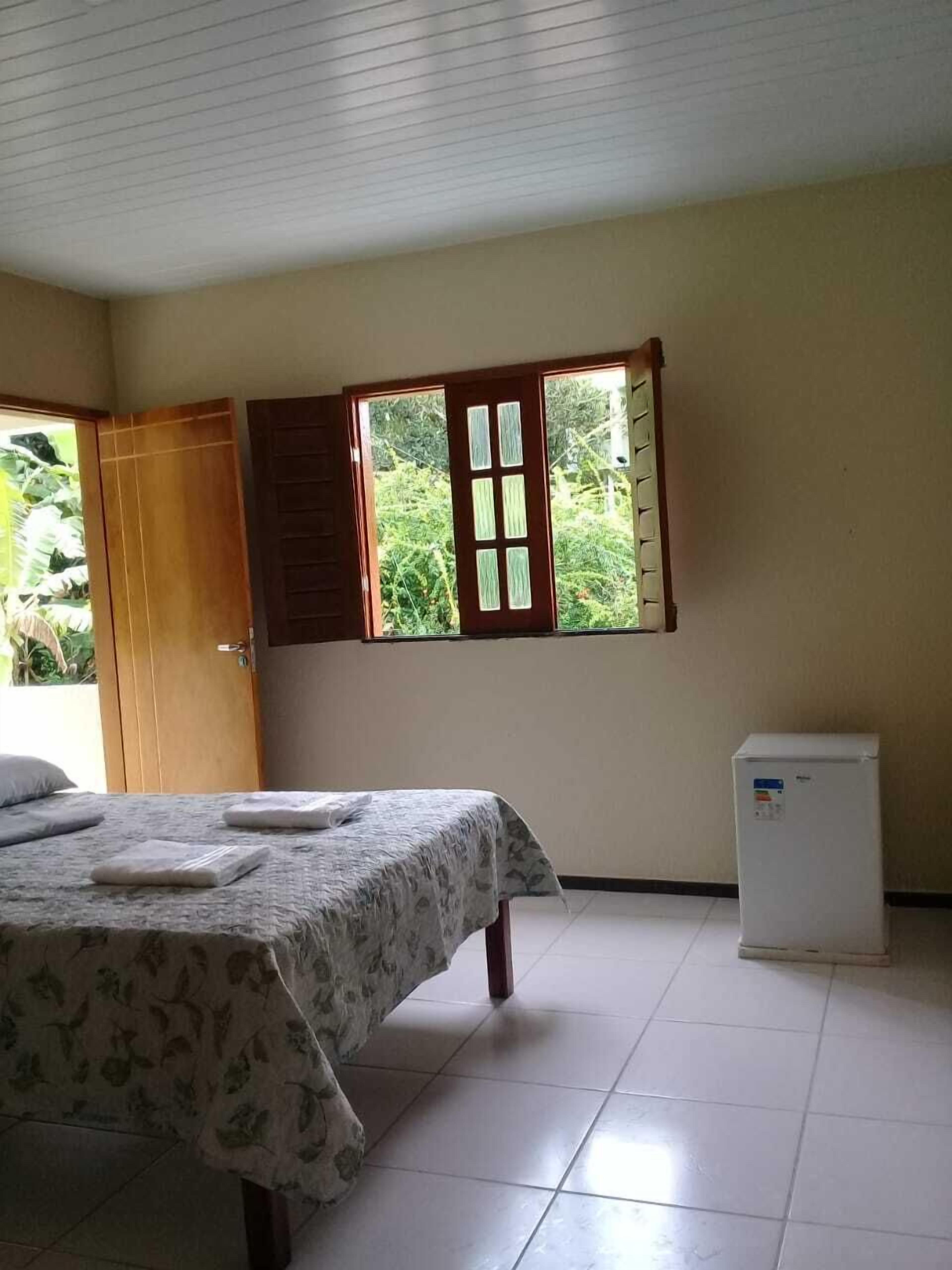A room with a bed, a window with wooden shutters, a small refrigerator, and a door leading outside.