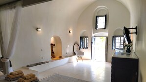 A spacious room with a bed, a desk, and a chair. There are two windows with open shutters, a wall-mounted TV, and a decorative sculpture.