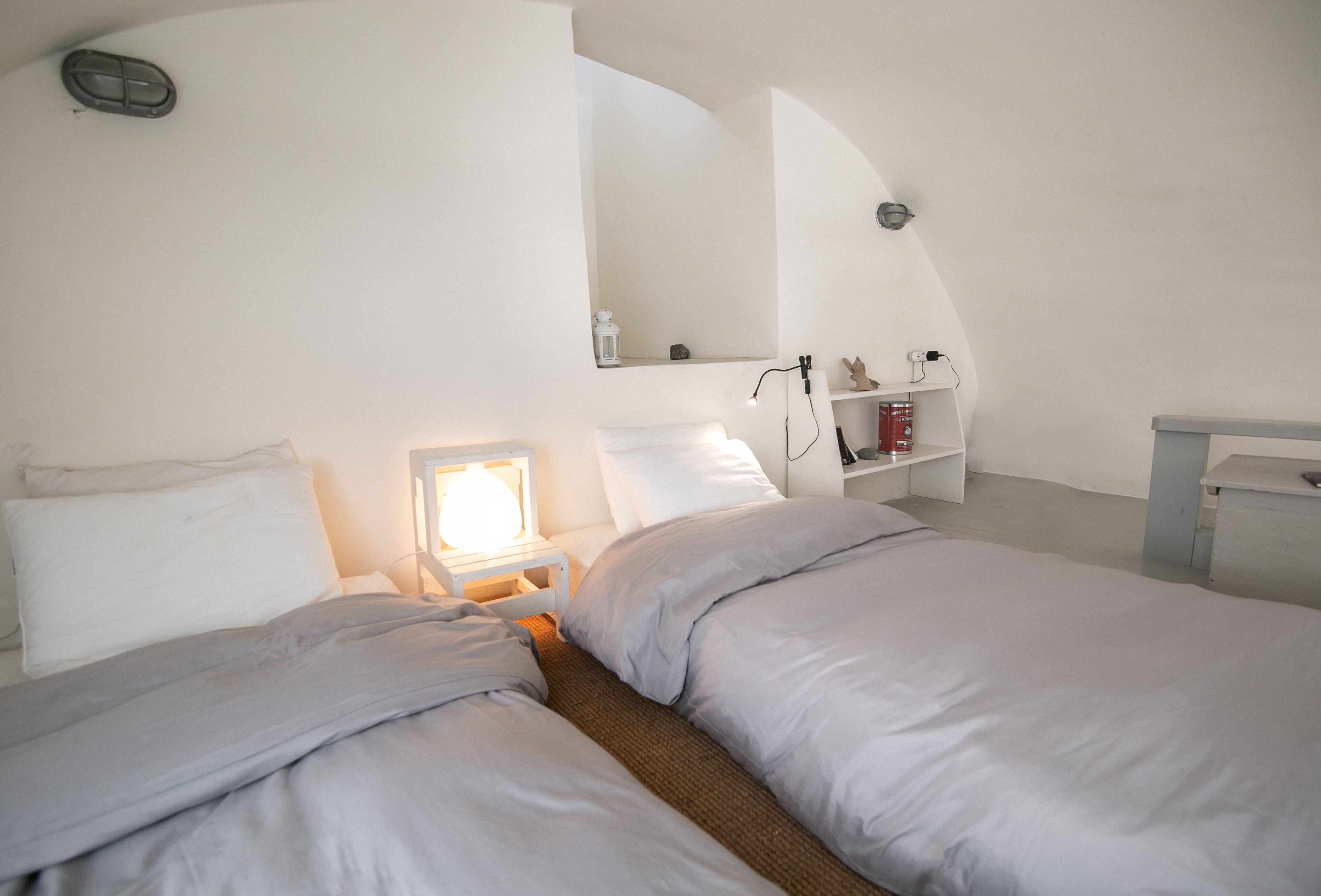 A bedroom with two beds, a bedside table with a lamp, a shelf with books and a small statue, and a mirror on the wall.