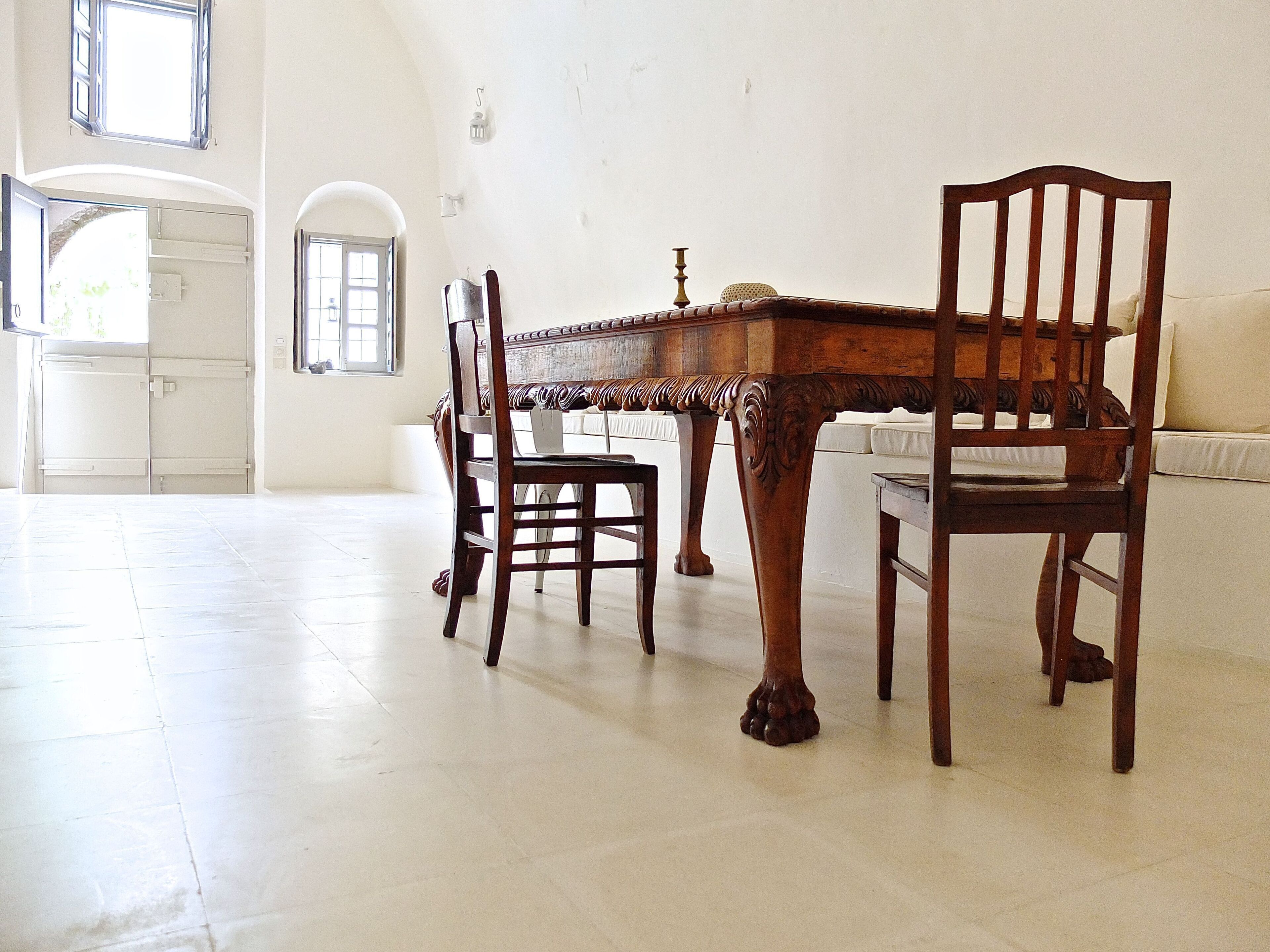 A dining area with a wooden table and chairs, a white tiled floor, and a white wall with a window.
