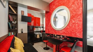 A modern hotel room with a red accent wall, a round mirror, a black dining table with red chairs, a black sofa with red cushions, and a kitchenette in the background. - ★CENTRAL LUXURY MINI SUITE JULIET★JUST RENOVATED (Θεσσαλονίκη)