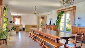 Un comedor amplio con una larga mesa de madera y bancos, rodeado de plantas en macetas y una araña.