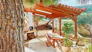 Una pérgola de madera con un muro de piedra y un hogar, rodeado de plantas en macetas y una mesa de piedra con sillas.