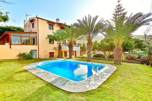 Una casa de dos pisos con piscina, rodeada de césped y palmeras.