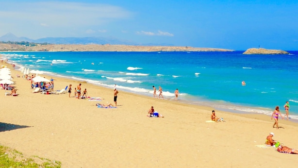 Una playa con gente tomando el sol y nadando, cielo despejado y montañas a lo lejos.