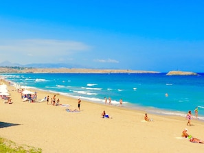 Una playa con gente tomando el sol y nadando, cielo despejado y montañas a lo lejos.