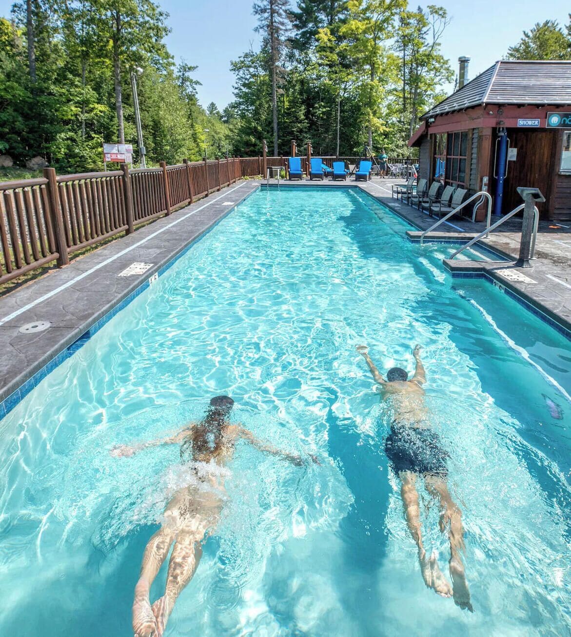 Une piscine où flottent deux personnes, entourée d’une terrasse en bois et d’une clôture.