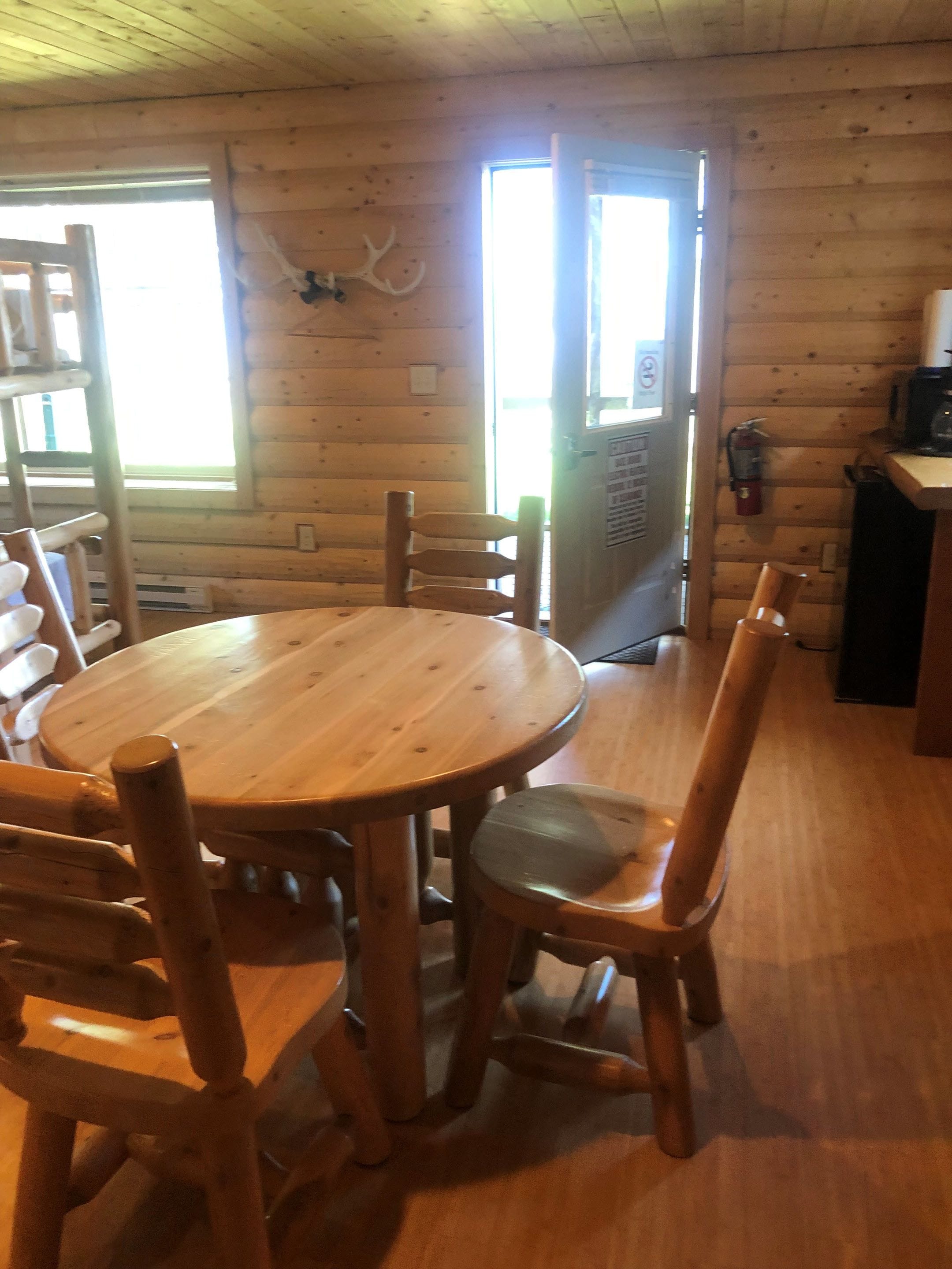 Un intérieur en bois avec une table ronde et des chaises, un extincteur et une porte donnant sur l’extérieur.