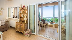 A living room with a brown sofa, a wooden cabinet, and a small table with a lamp. There is a sliding glass door leading to an outdoor patio with a chair and a table.