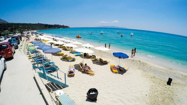 A beach with rows of sun loungers and umbrellas, a parking area, and boats in the water.