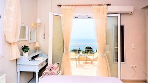 A bedroom with a bed, a desk, a chair, a potted plant, and a view of the sea through a balcony.