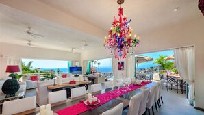 Une vaste salle à manger avec une longue table dressée pour le repas, un lustre décoratif et une vue sur l’océan.