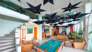 Un vaste espace de vie comprenant une table de billard, des sièges confortables, un escalier et, au fond, une cuisine moderne.