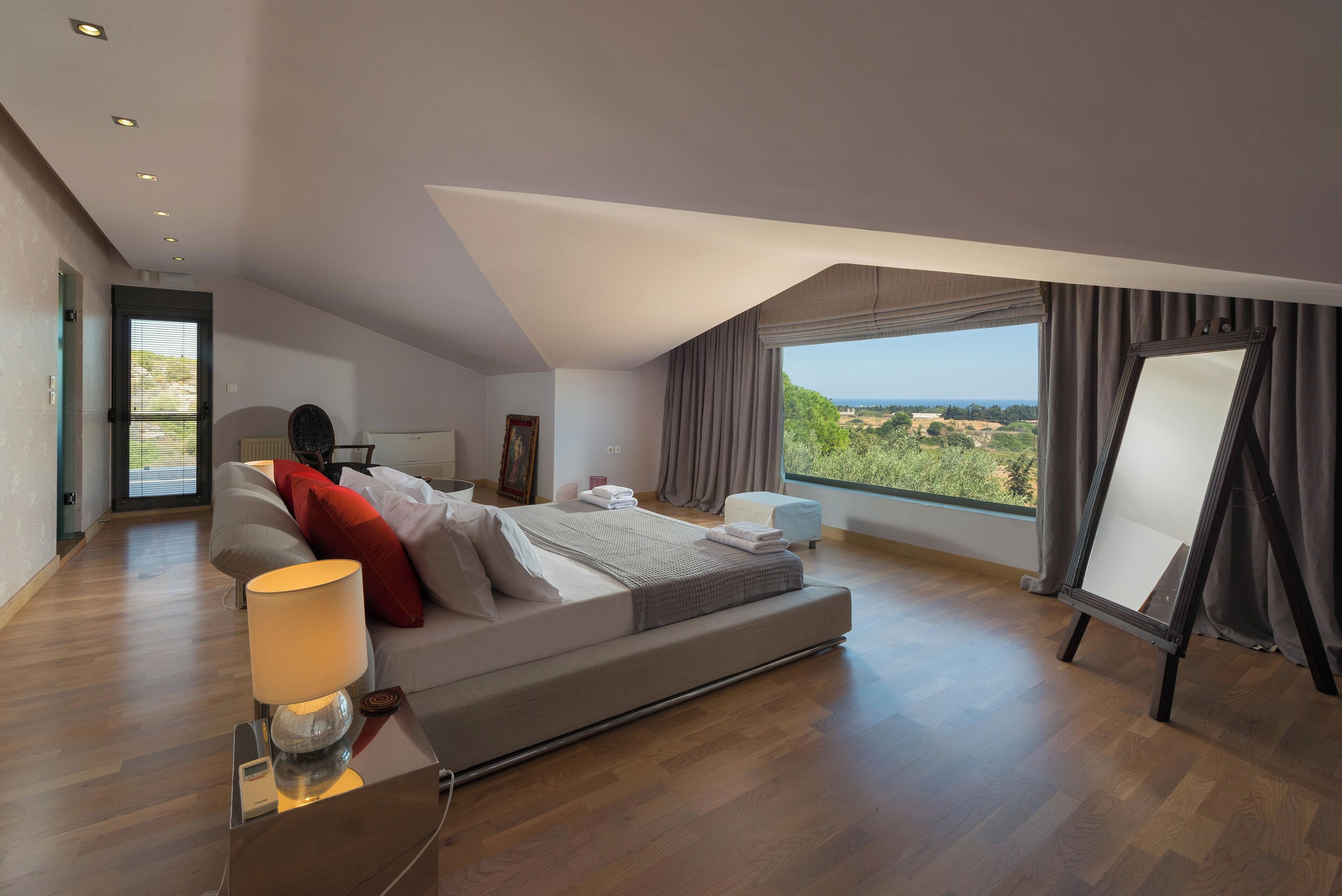 A modern bedroom with a large bed, a standing lamp, a view of the outdoors, and a painting on the wall.