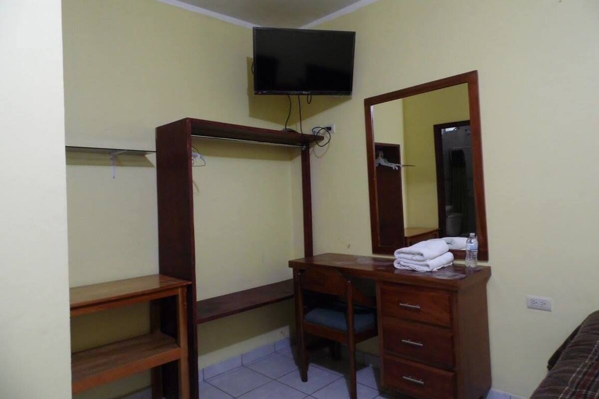 A room with a wooden desk, a chair, a television mounted on the wall, and a mirror above the desk.