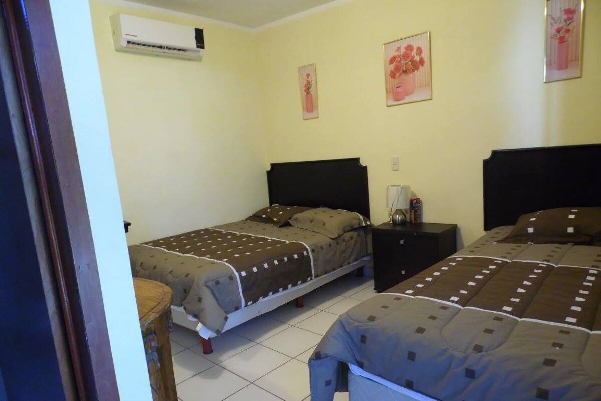 A room with two beds, a bedside table, an air conditioner, and two framed pictures on the wall.