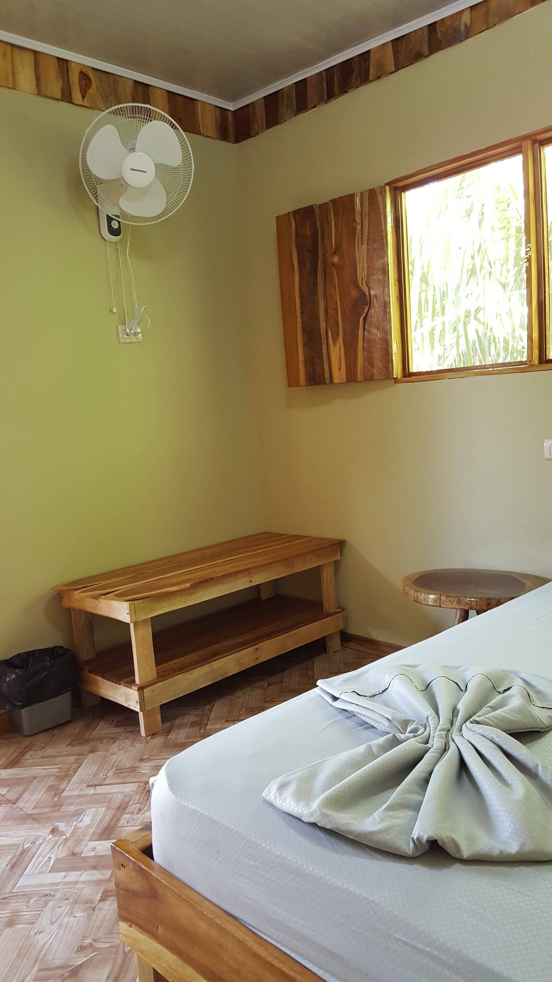 Une pièce avec un banc en bois, un lit avec une couverture soigneusement pliée, un ventilateur sur pied et une fenêtre donnant sur un paysage verdoyant.
