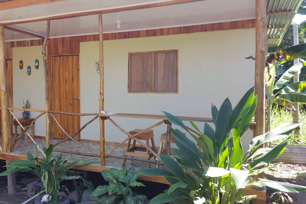 A wooden porch with a simple railing, a door, and a window. There are potted plants around the porch area.
