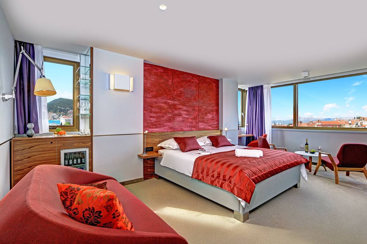 A hotel room with a bed, a sofa, a small table, and a chair. There is a window with a view of buildings and a mountain.