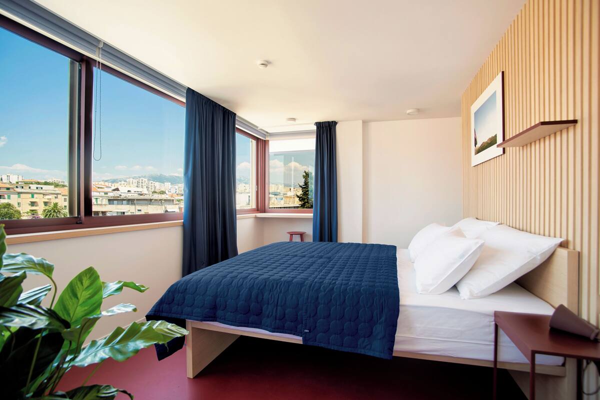 A modern hotel room with a large bed, a small stool, a window with curtains, and a city view.