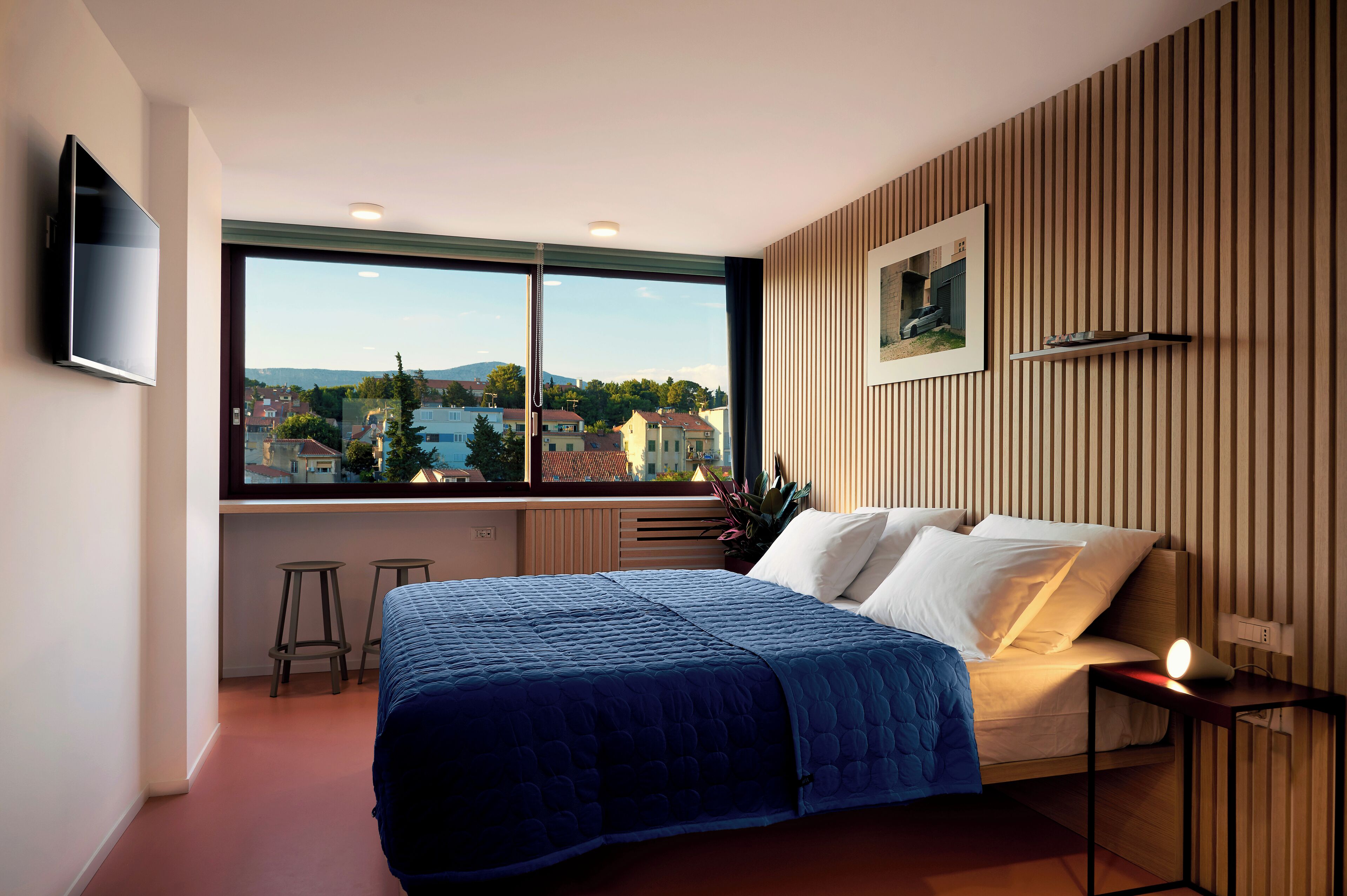Un dormitorio con un ventanal grande, una cama, un televisor y una mesita.