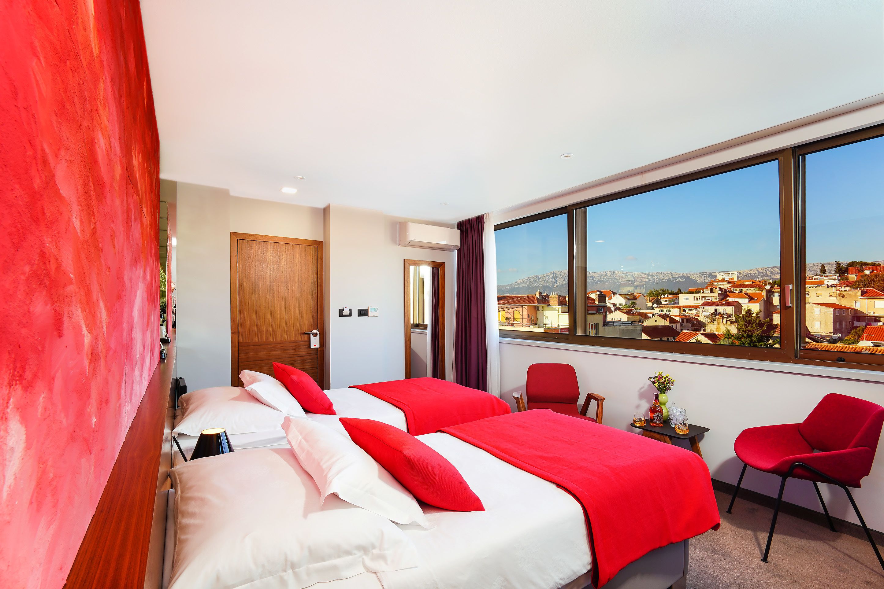 Une chambre d’hôtel avec deux lits, une grande fenêtre, un fauteuil rouge et une vue sur la ville.