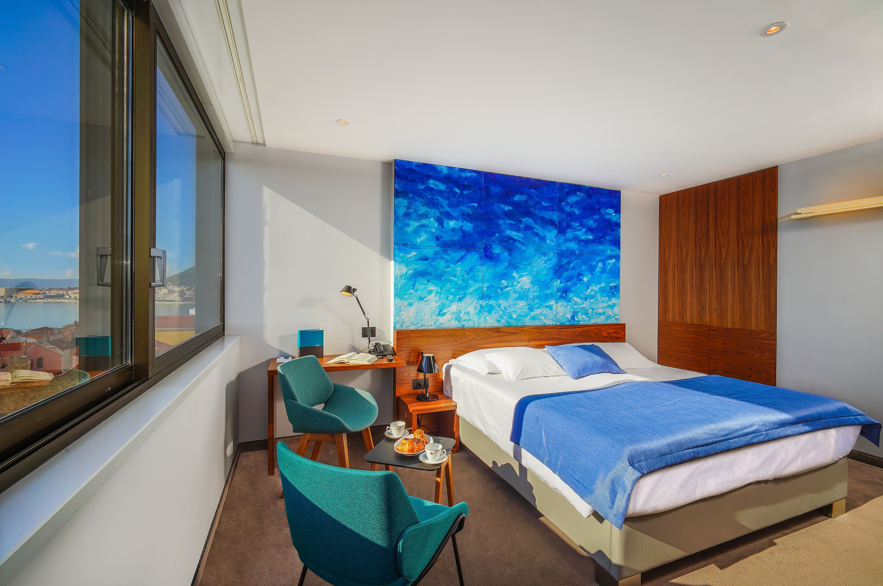 A hotel room with a large window, a bed with blue bedding, a desk with a lamp, and a chair.