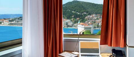 A room with a desk, laptop, and phone, overlooking a coastal town and hills.