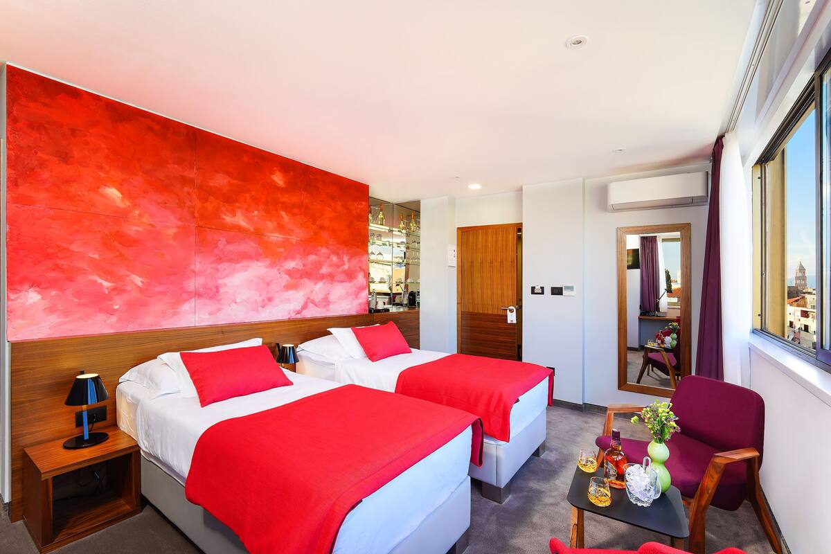 A hotel room with two beds, a wooden headboard, a red accent wall, a small table with a vase and drinks, and a window with a view.