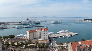 Un paisaje urbano costero con un edificio destacado que tiene una cúpula, un puerto deportivo con varios cruceros, y una vista despejada del mar y la costa lejana.