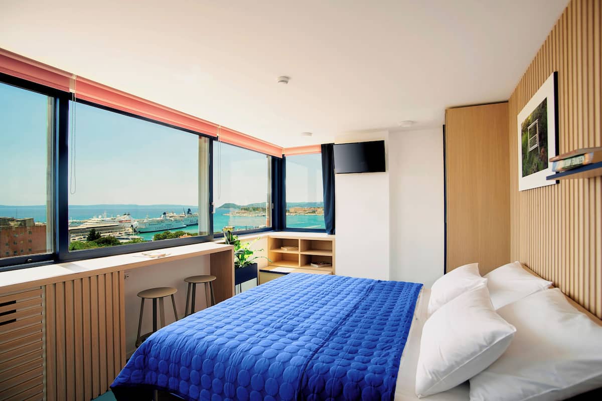 A bedroom with a large window, a bed with white pillows, a blue bedspread, a wooden desk, and a TV on the wall.