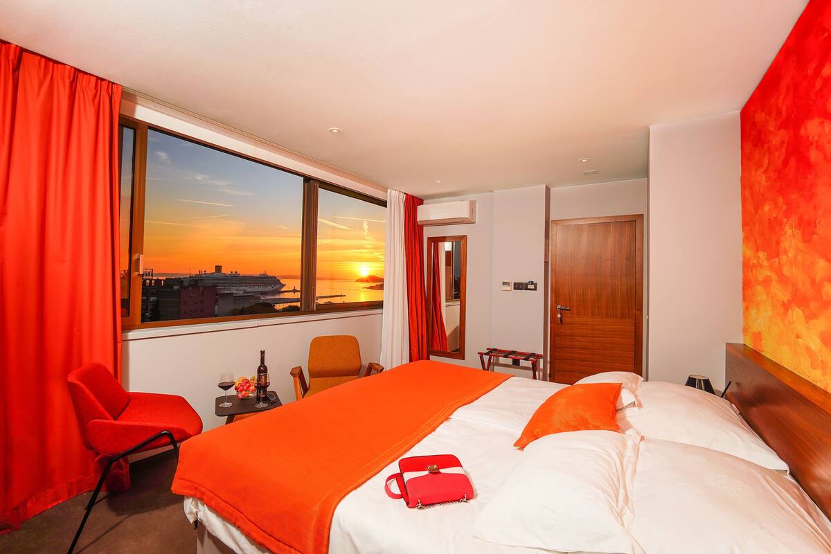 A hotel room with a large bed, a chair, a small table with a bottle and glasses, and a view of the sunset through a large window.