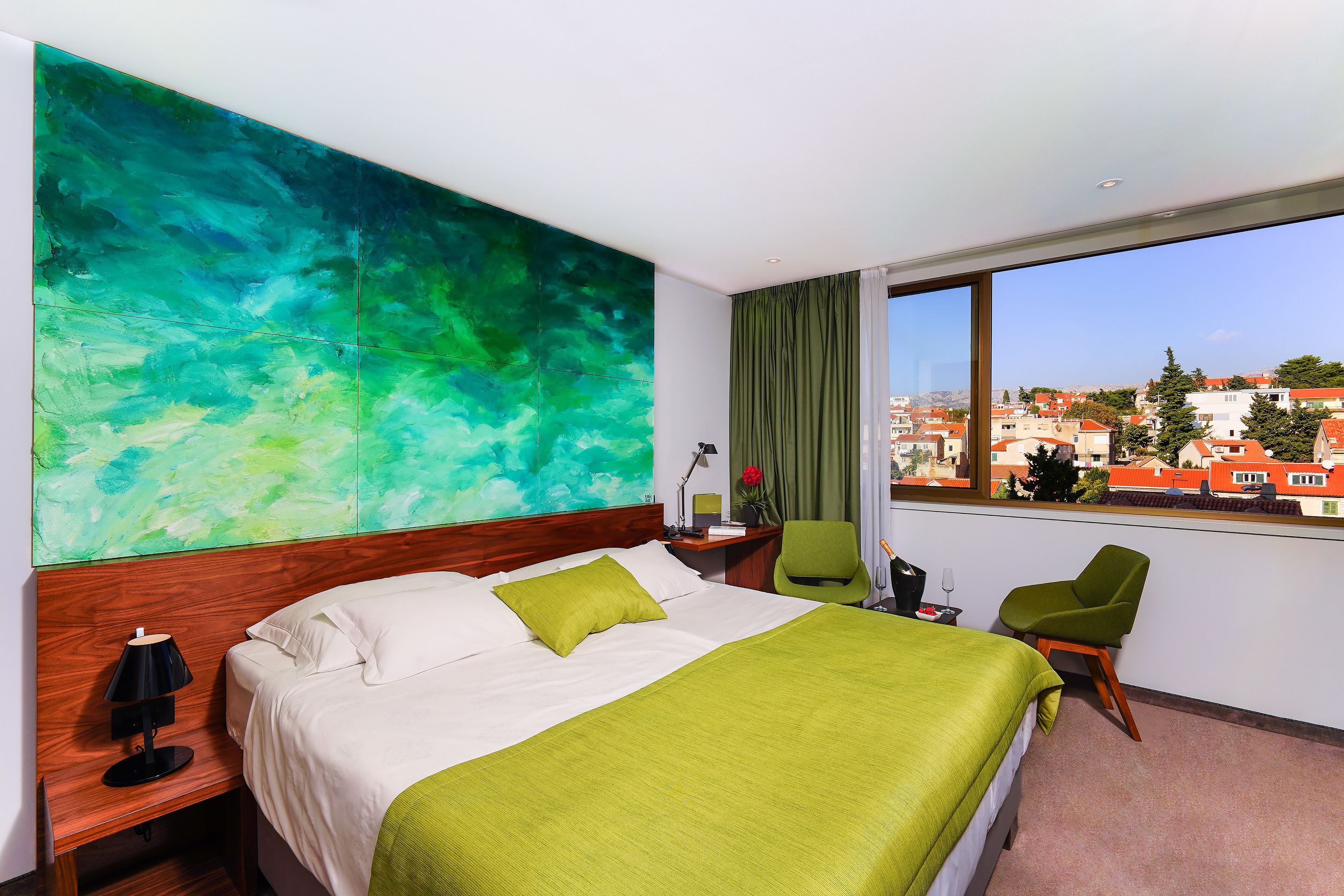 A hotel room with a large bed, green and white bedding, a green chair, a desk with a lamp, and a view of a cityscape through a large window.