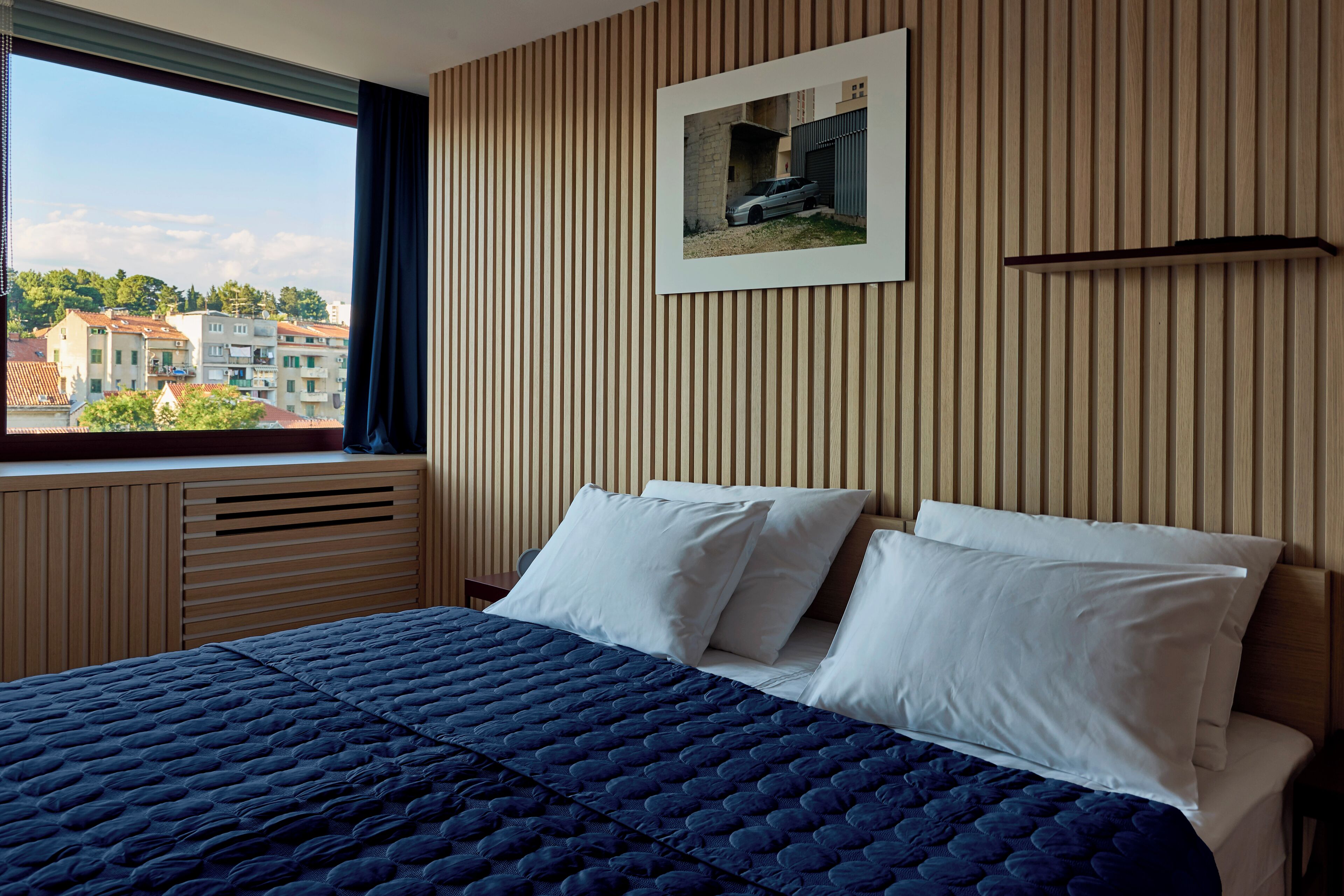 A bedroom with a large bed, a window with curtains, a wall-mounted picture, and a view of buildings outside.